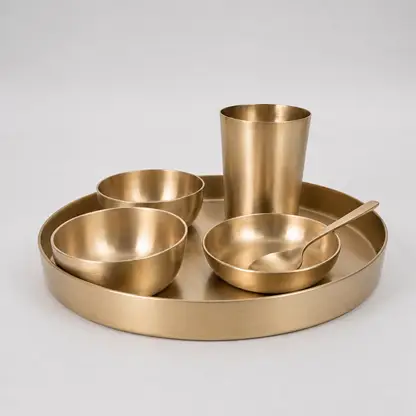A set of matte brass tableware including a round tray, a tumbler, two bowls, and a spoon, arranged on a light gray background.