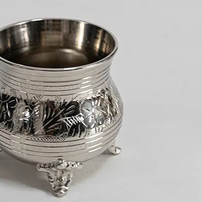 Closeup of silver teapot with ornate detailing curved spout lid and handle on footed base placed on white background.