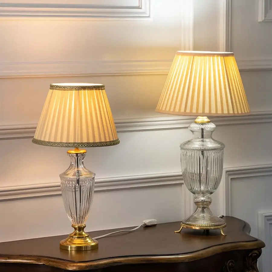 Pair of crystal glass table lamps with gold bases and pleated beige shades turned on together adding soft warm illumination to classic home interiors.