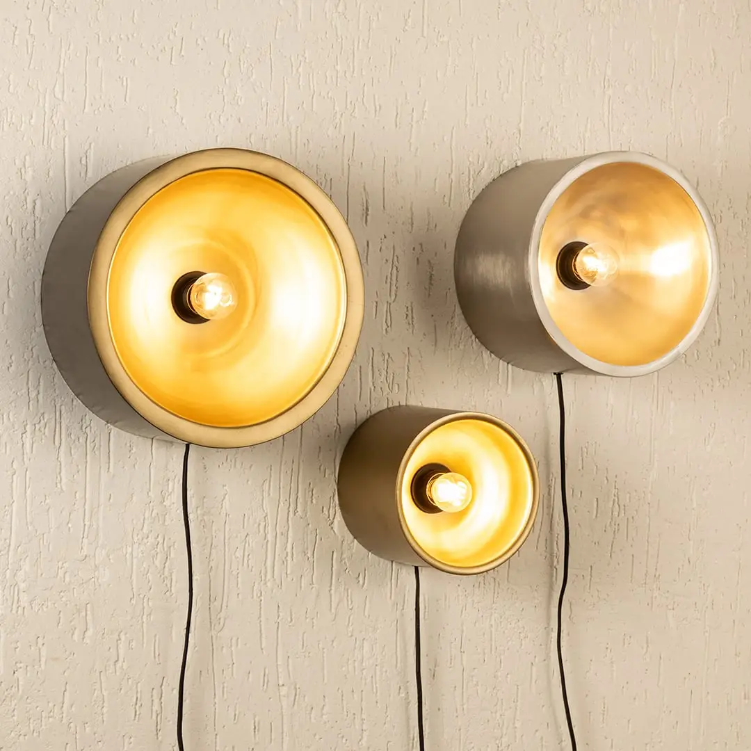 Trio of modern gold and silver cylinder cup wall sconce lamps casting a warm glow clustered on a textured beige wall.