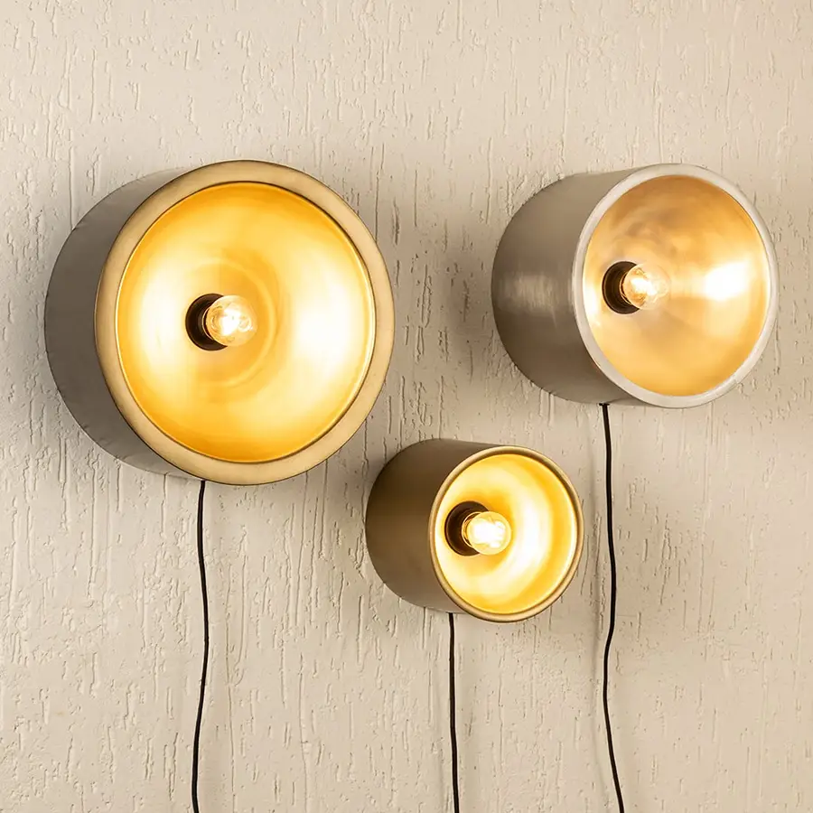 Trio of modern gold and silver cylinder cup wall sconce lamps casting a warm glow clustered on a textured beige wall.