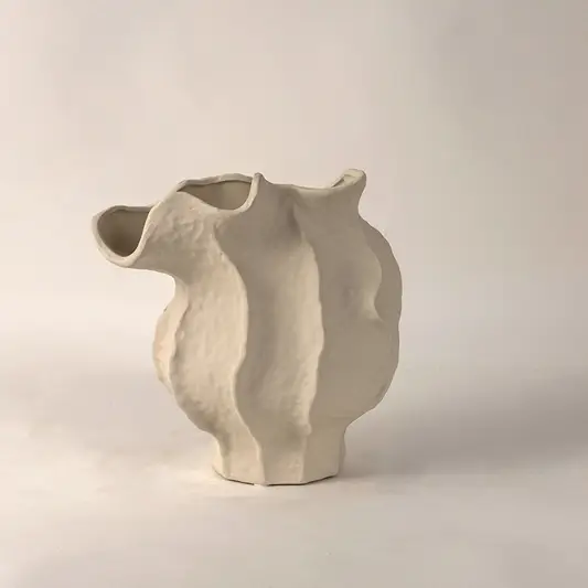 White ceramic vase with irregular curved sculptural form placed on white surface against neutral background highlighting contemporary artistic home decor appeal.