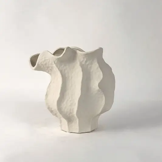 White ceramic sculptural vase with organic flowing shape placed on white surface against neutral background showcasing elegant modern home decor styling.