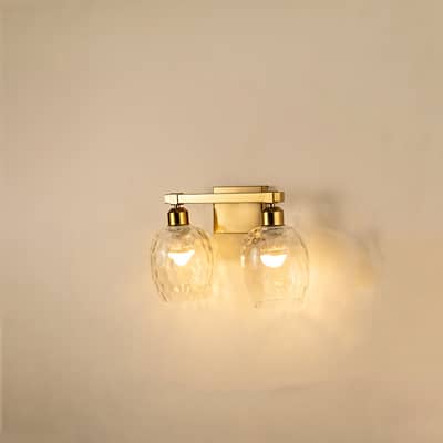 Brushed gold double wall light with hammered clear glass shades glowing on white background adding warm elegant lighting to modern interiors.