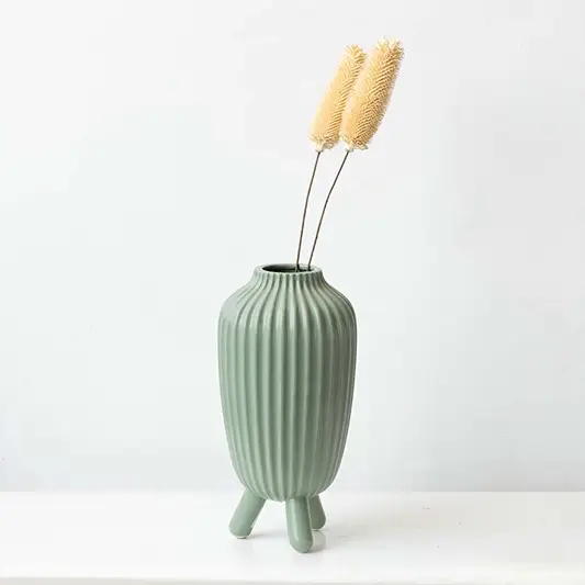 Two natural cream beige dried cattail bulrush stems arranged inside a modern ribbed green ceramic vase on white background.