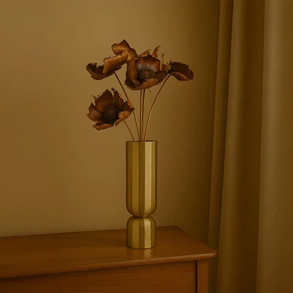 A tall gold metal vase holding dried brown flowers placed on a wooden surface against a warm beige backdrop creating elegant earthy botanical accents for sophisticated interior décor.