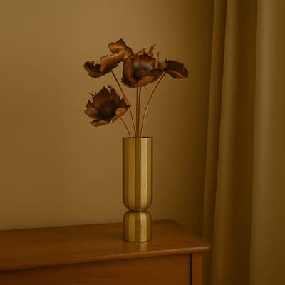 A tall gold metal vase holding dried brown flowers placed on a wooden surface against a warm beige backdrop creating elegant earthy botanical accents for sophisticated interior décor.