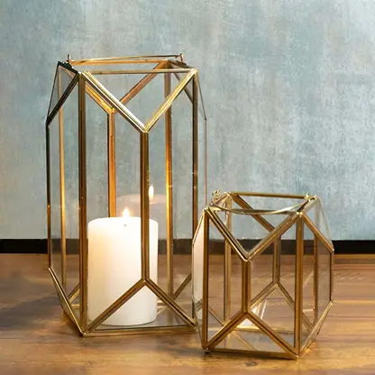 Two gold metal and glass candle lanterns grouped together with candles on creating a warm decorative display perfect for tabletops floors or corners.