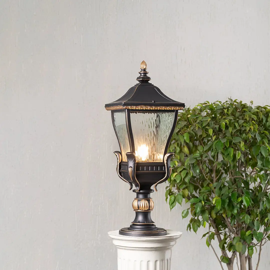 Elegant black and gold metal outdoor pillar light glowing with clear glass shade placed on decorative column lifestyle view.