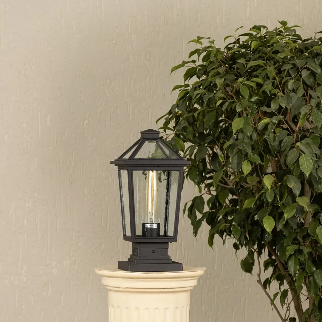 Black outdoor pillar light with clear glass panels placed by a large green plant Lifestyle image patio decor.