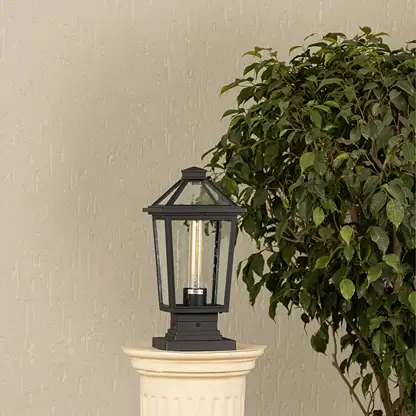 Black outdoor pillar light with clear glass panels placed by a large green plant Lifestyle image patio decor.