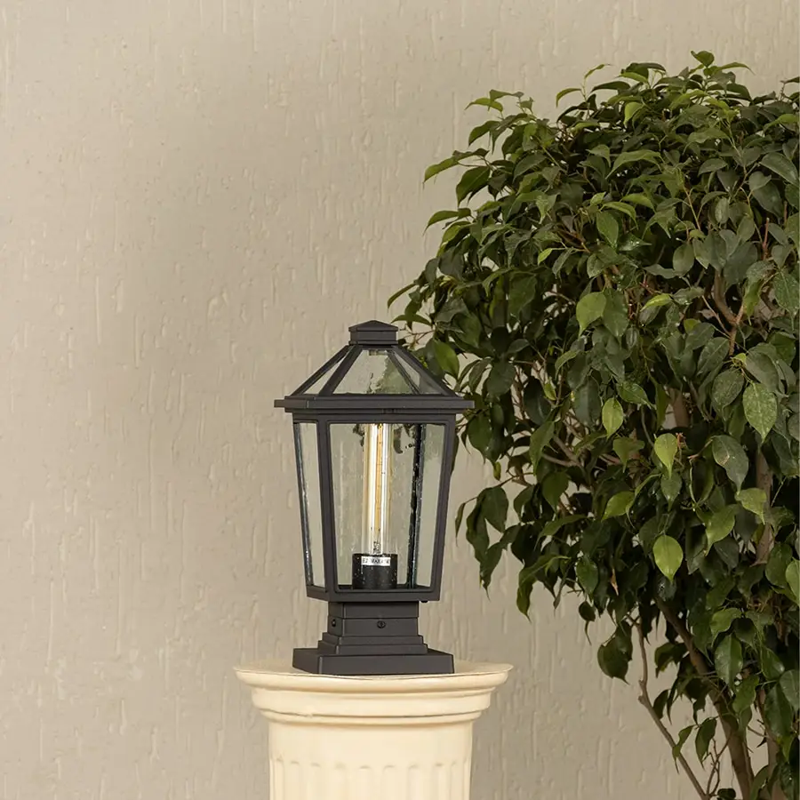 Black outdoor pillar light with clear glass panels placed by a large green plant Lifestyle image patio decor.