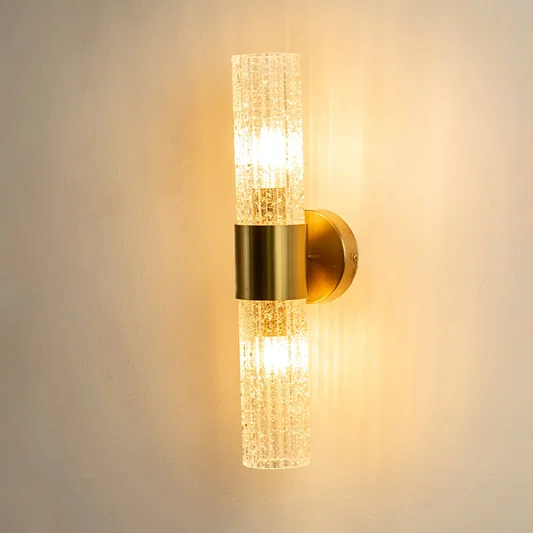 A gold wall sconce with two textured cylindrical glass shades shown with lights on against a white background highlighting its warm ambient glow and vertical design.