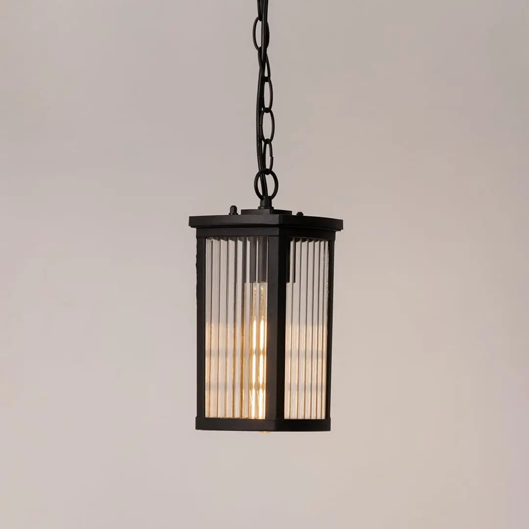 Black metal hanging lantern lit on white background highlighting warm glow and ribbed glass for home lighting.