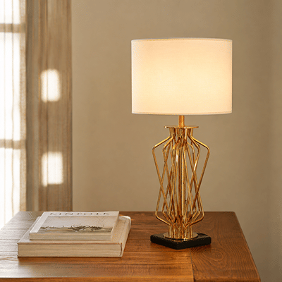 A modern table lamp with a glowing white drum shade and a gold geometric wireframe base on a black square pedestal, set on a wooden table next to stacked books.
