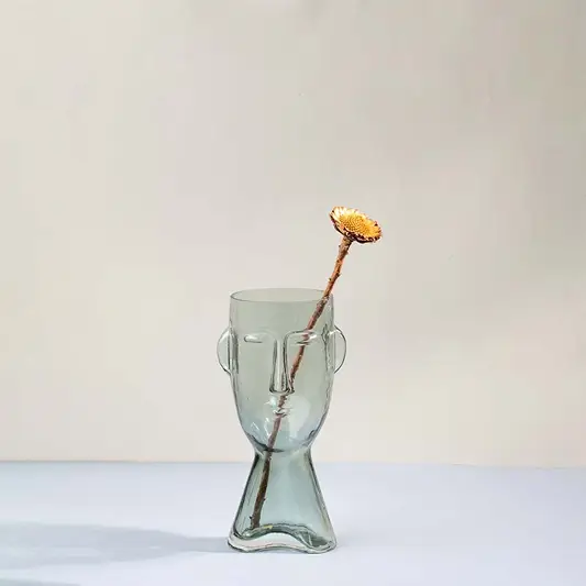 Modern glass vase in abstract face design transparent finish styled with single dry flower for artistic living room placement.