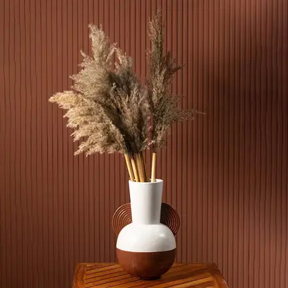 Pampas grass stems displayed in a modern ceramic vase with brown and white finish placed on a wooden table against a terracotta wall.