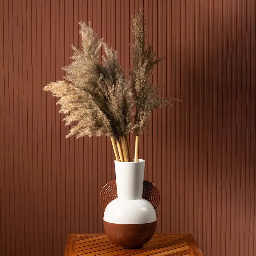 Pampas grass stems displayed in a modern ceramic vase with brown and white finish placed on a wooden table against a terracotta wall.