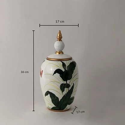 White ceramic decor jar with tropical green leaf artwork and golden lid shown with 36 cm height and 17 cm width.