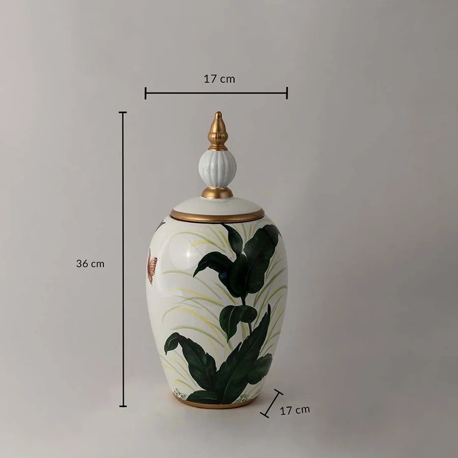 White ceramic decor jar with tropical green leaf artwork and golden lid shown with 36 cm height and 17 cm width.