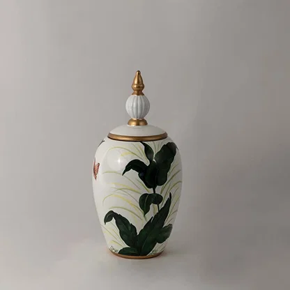 Decorative ceramic jar with green botanical leaf artwork and gold lid placed on white surface against neutral background showcasing elegant modern home decor styling.