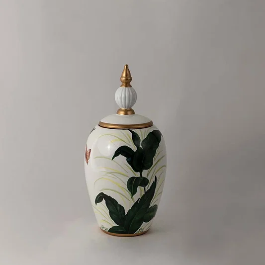Decorative ceramic jar with green botanical leaf artwork and gold lid placed on white surface against neutral background showcasing elegant modern home decor styling.