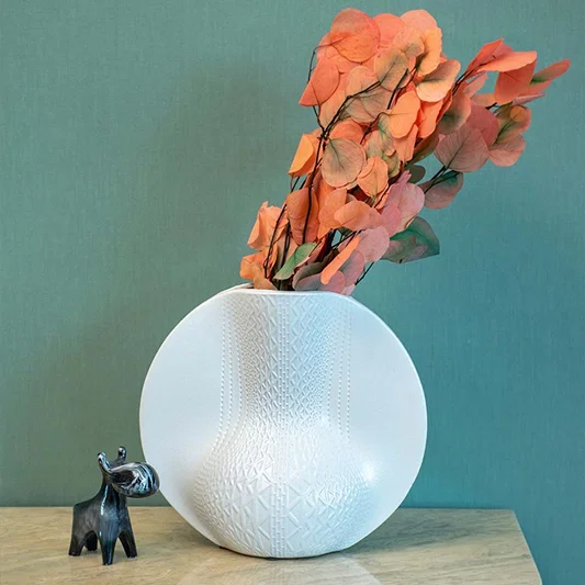 Round white pottery vase with geometric pattern placed on a stone surface against a teal wall for contemporary home decor styling.