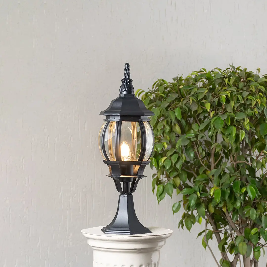 Black outdoor pillar lamp glowing warmly with LED bulb and clear curved glass shade perfect for patio driveway and terrace.