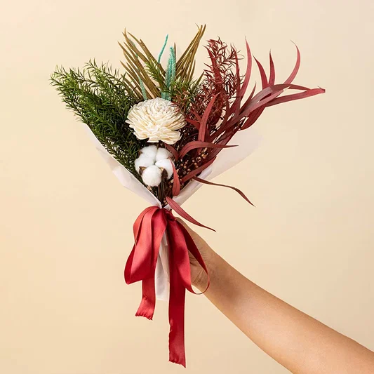 Hand holding a rustic dried floral bouquet with white cotton pods and red preserved leaves tied with a red satin ribbon.