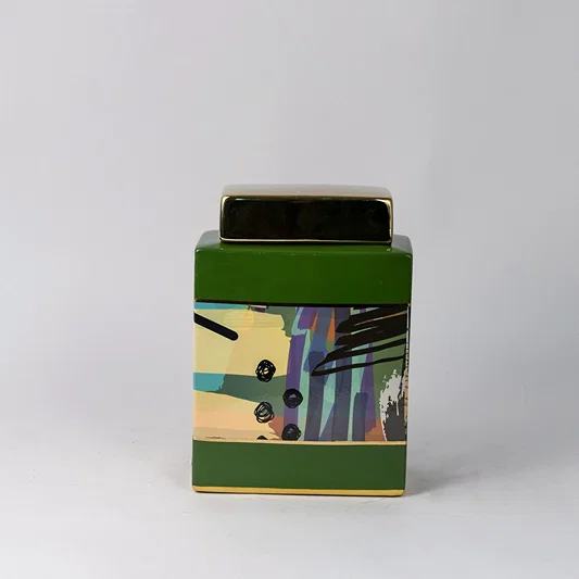 Square ceramic jar with green base and abstract artwork placed on white surface against neutral background showcasing contemporary modern home decor styling.