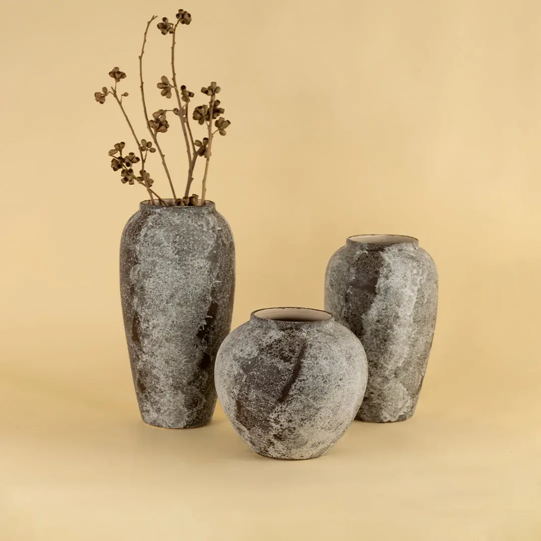 Set of grey textured ceramic vases with dried stems placed on beige surface against warm background showcasing rustic modern home decor styling.