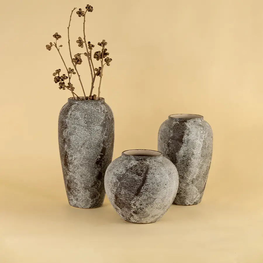 Set of grey textured ceramic vases with dried stems placed on beige surface against warm background showcasing rustic modern home decor styling.