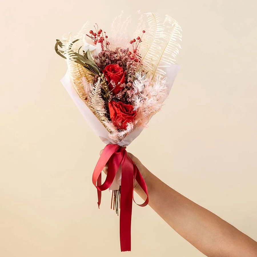 Hand holding a classic red roses dried flower bouquet tied with a red ribbon for luxury romantic anniversary gifting.