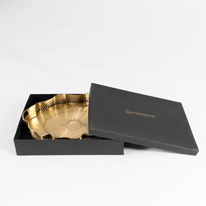 Elegant scalloped brass tray partially placed inside black gift box with gold logo shown on white surface.