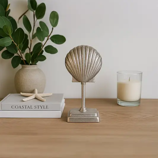 A decorative seashell figurine on a stand displayed on a wooden table with a candle green leaves and coastal themed books against a white background.