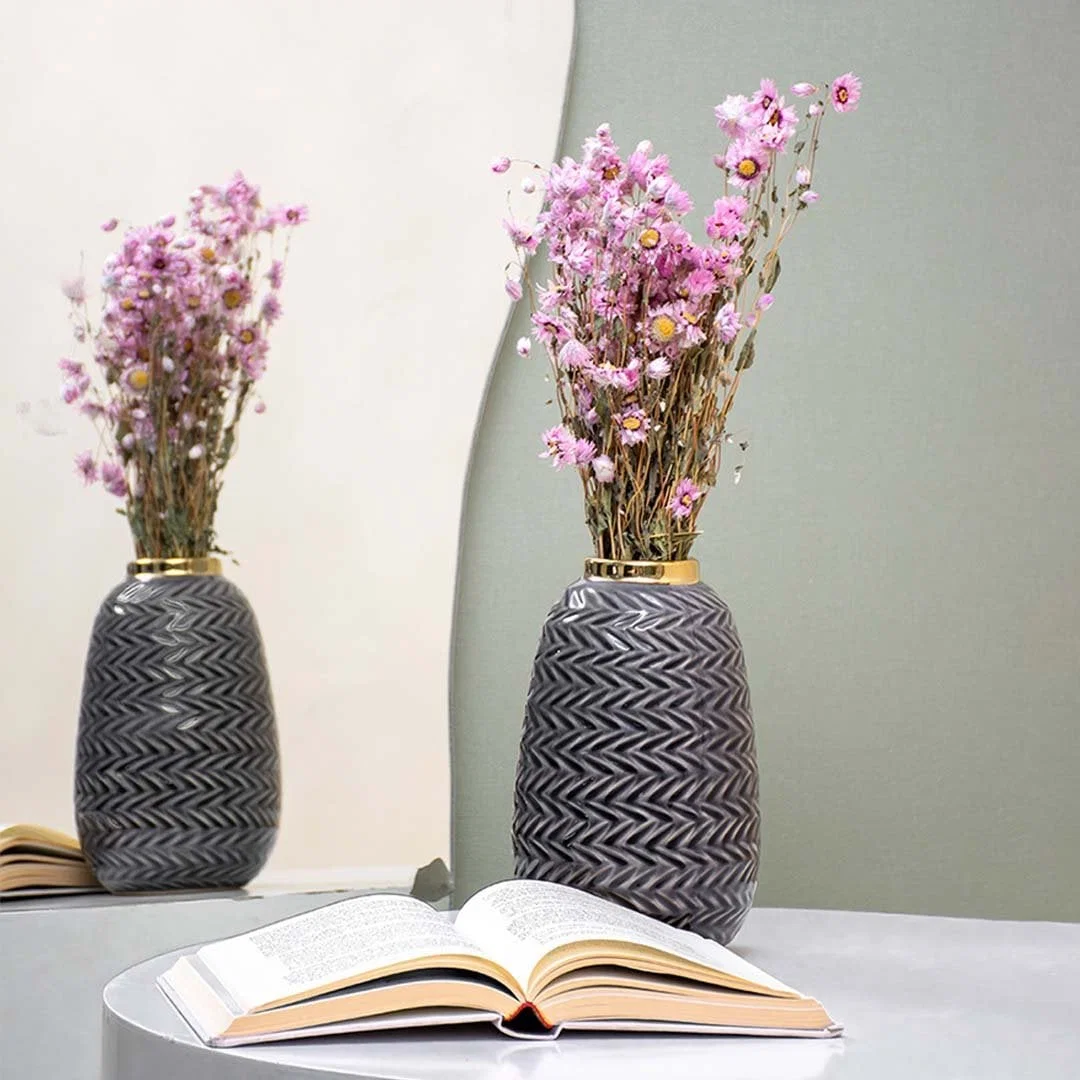 A decorative grey vase with a zigzag pattern filled with pink dried flowers placed beside an open book on a reflective table creating a soft elegant lifestyle scene.