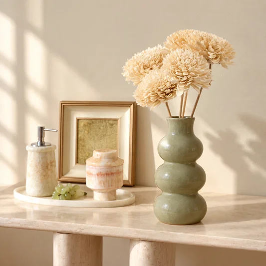A three-bottle ceramic vase in a muted sage green glaze holding dried cream chrysanthemums, styled on a light stone console table beside a soap dispenser, small jar, and framed picture.