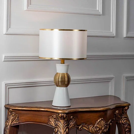 Table lamp with white ceramic base and gold accents with white fabric shade on wooden table modern classic lighting for decor.
