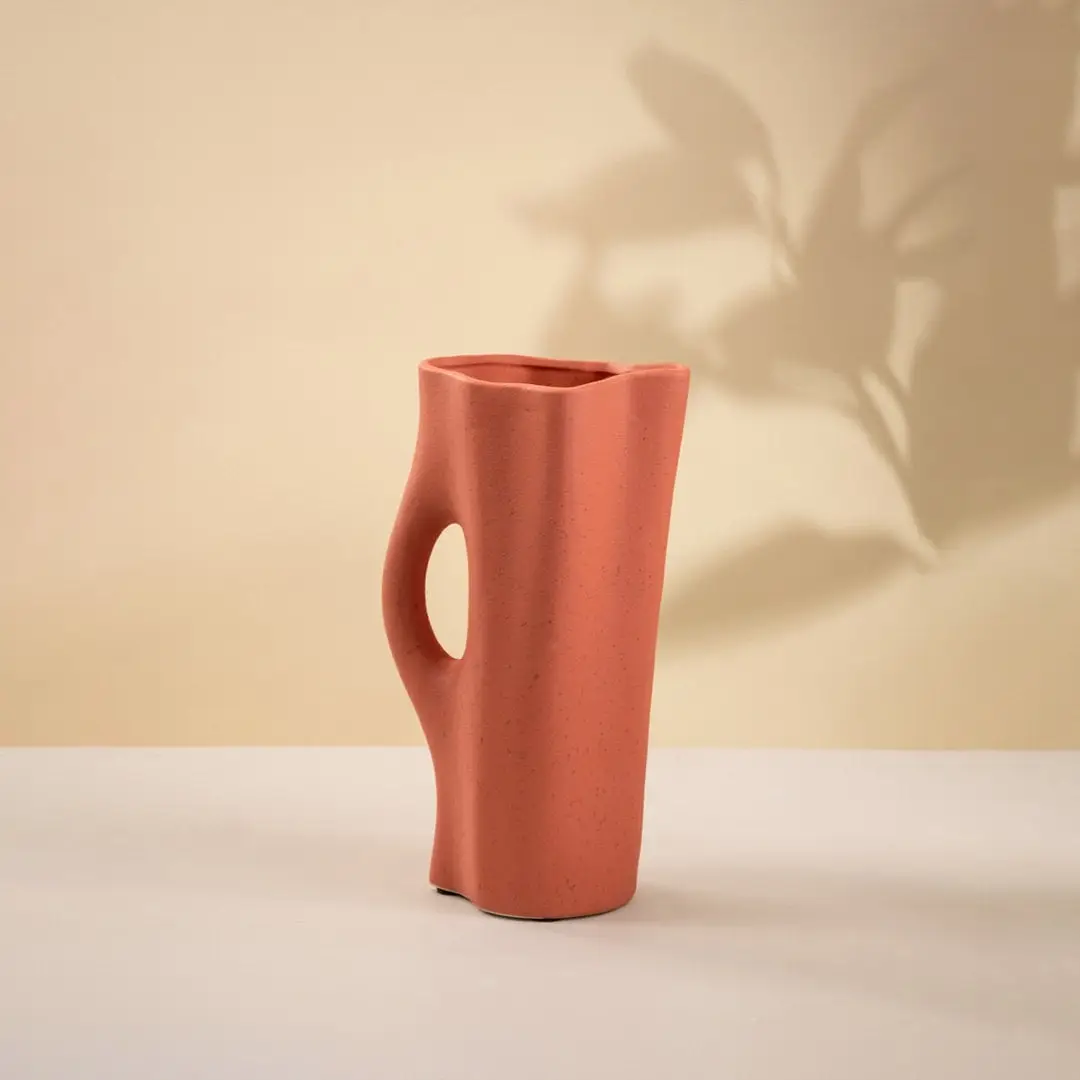 Contemporary terracotta ceramic vase with matte surface and sculpted handle-style cutout placed on a plain white background.
