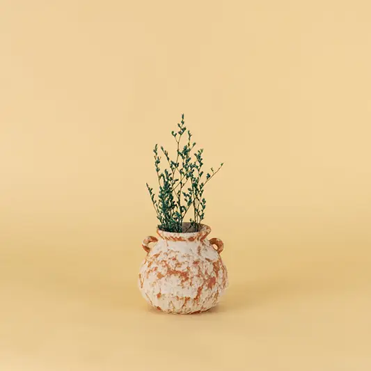 Small ceramic vase with floral artwork and green plant stems placed on beige surface against warm background enhancing minimal botanical home decor aesthetic.