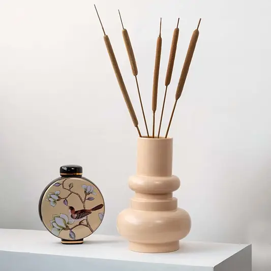 Brown cattail stems styled in beige ceramic vase placed on modern table for elegant home decoration.