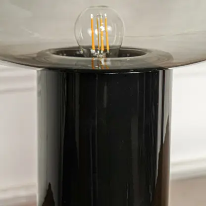 Close up black metal base table lamp with exposed filament bulb and glass shade positioned on wooden surface against neutral wall background.