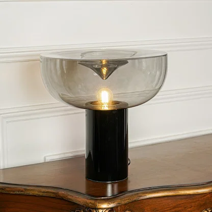 Elegant tinted glass table lamp with gold metal stem and black base positioned on wooden surface against minimal wall background for ambient lighting.
