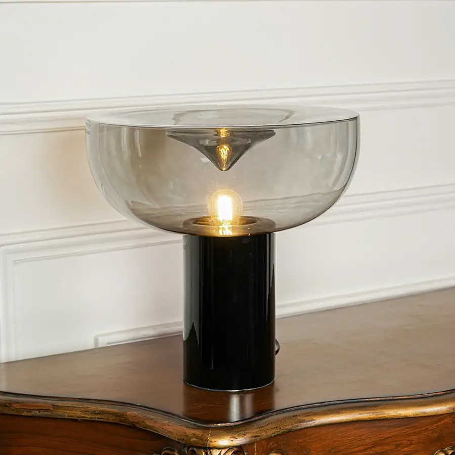 Elegant tinted glass table lamp with gold metal stem and black base positioned on wooden surface against minimal wall background for ambient lighting.
