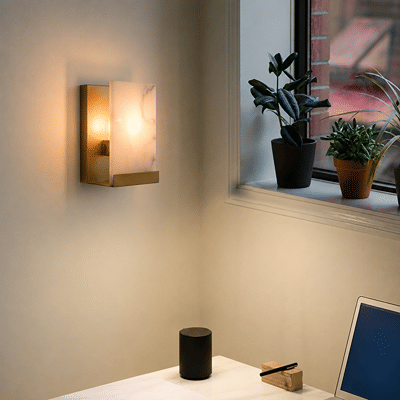A modern wall-mounted sconce with a rectangular gold-toned metal frame and a glowing warm white marble-look glass shade, installed on a beige wall in a room setting next to a window with potted plants.