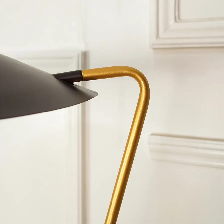 Close-up of a modern floor lamp with a matte black conical shade and a curved gold metal stem, set against a white paneled wall.