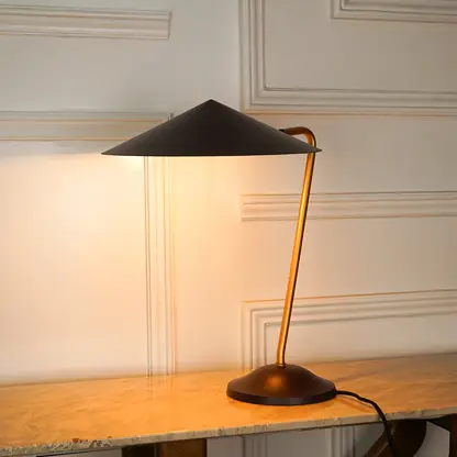 A modern table lamp with a black conical shade, a curved gold-toned metal stem, and a round dark base, illuminated and placed on a light wood console table against an off-white paneled wall.