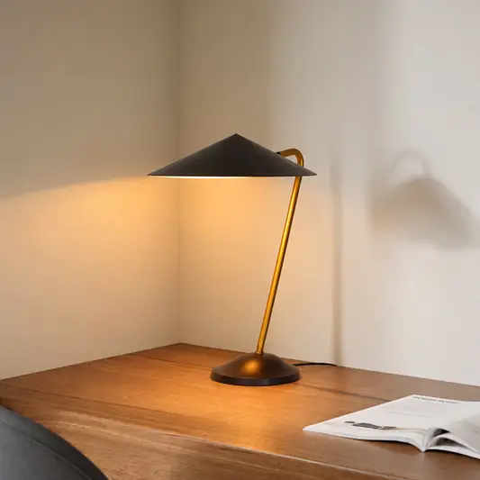 A modern table lamp with a matte black conical shade and a warm brass-colored curved stem, illuminated on a wooden desk against a beige wall.