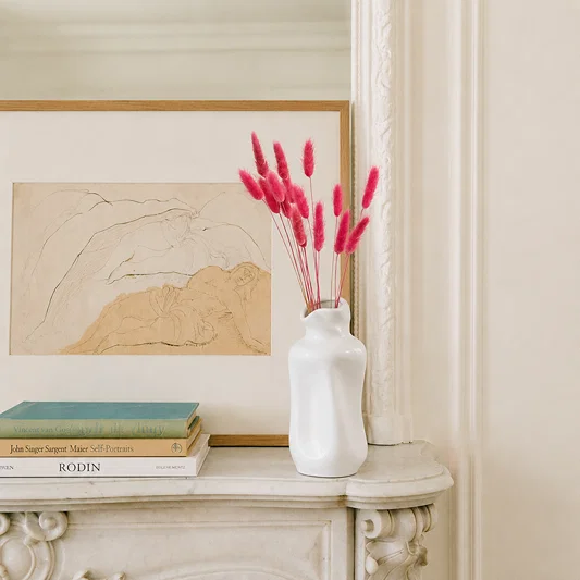 A white ceramic vase filled with bright pink bunny tail grass stems sits on a cream-colored mantelpiece next to a framed abstract landscape print and stacked books.
