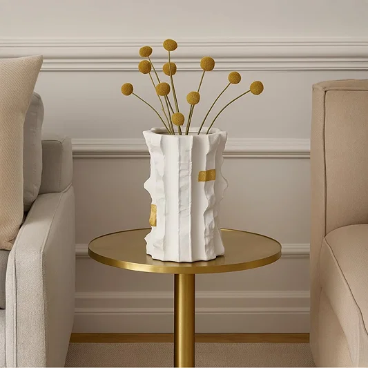 Astra white ceramic vase with gold accent details styled on a round brass table between two beige sofas adding a modern luxury touch to living room decor.